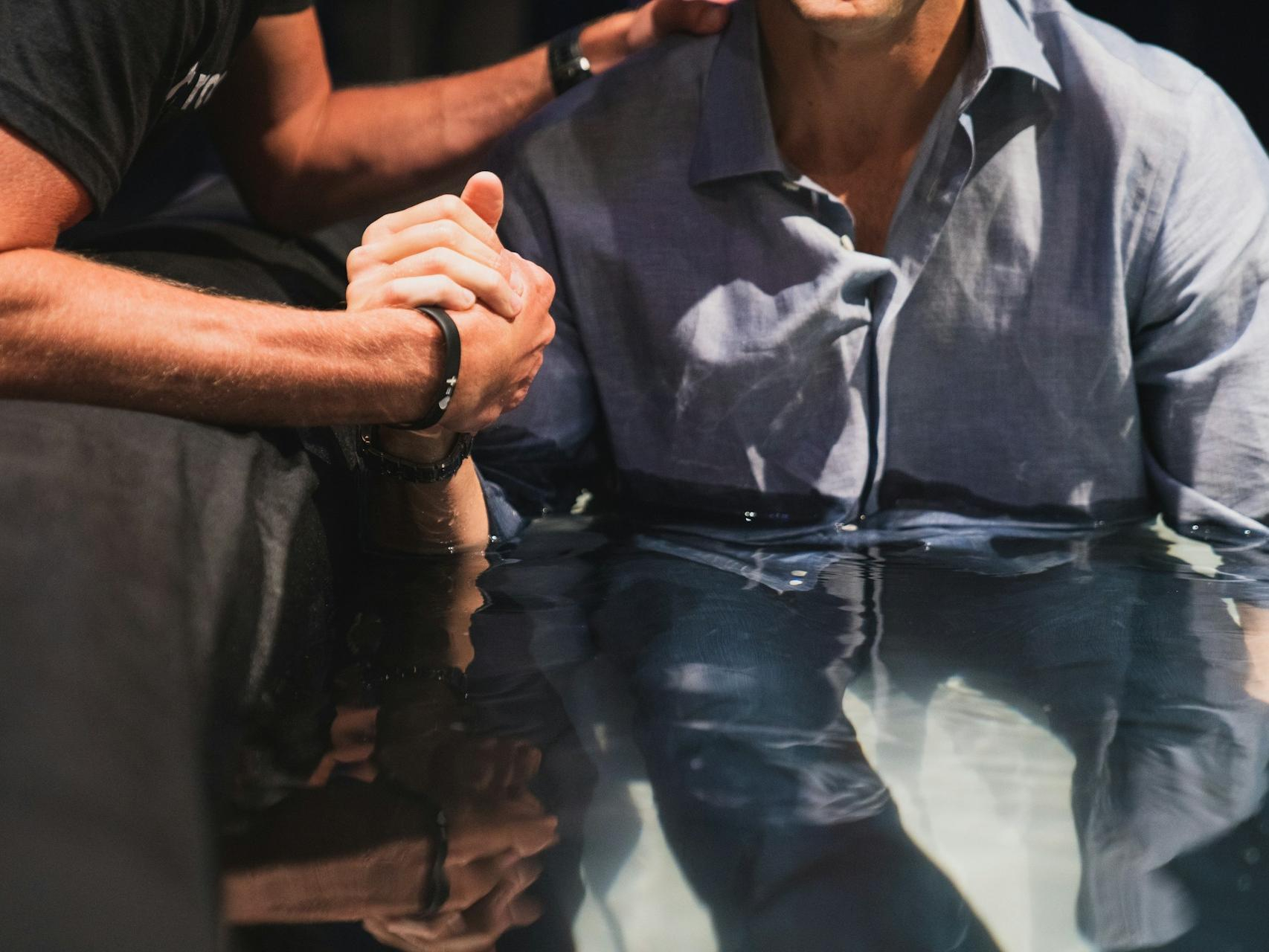 Ein Mann im Blauen Hemd wird getauft im Wasser und hält die Hand des Pastors der ihn eine Hand auf die Schulter legt