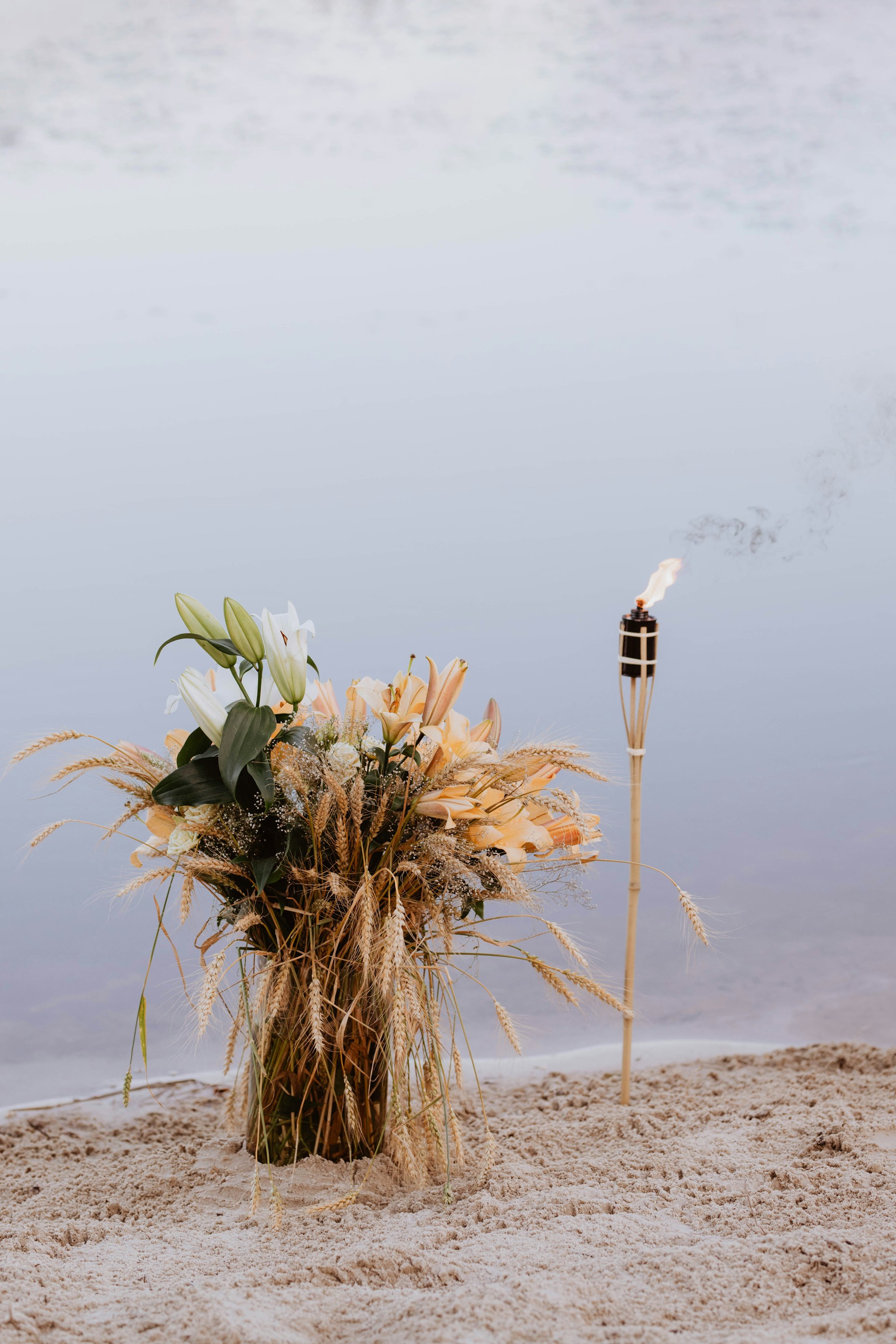 Ufer eines Sees mit einer Fackel die im Sand steckt und einem Strauß aus Weizen und Grüne Pflanzen